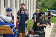 Students with suitcases, pillows and boxes walk next to a columned building on a college campus.