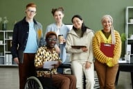 A small, diverse group of student leaders smiles at the camera in an office space. One is a wheelchair user. They have folders and coffee, as if in a meeting.