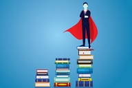 Man in a suit and a superman cape standing on a stack of books with two other stacks to the side of him