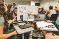 Close-up shot of four female students doing calculus problems on a whiteboard in a larger active learning–style classroom.