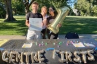 Three students stand at a black table with golden balloon letters that read "Centre Firsts"