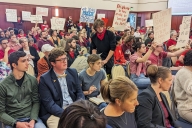 A photo of West Virginia University students, faculty members and others sitting and standing in a room at West Virginia University, with some holding signs protesting proposed academic program and faculty position cuts.