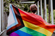 A young person carries a Progress Pride flag unfurled behind their back. 