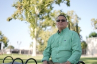 Daymon Johnson, a light-skinned man with dark hair wearing sunglasses and a long-sleeved green button-down shirt, sits on a bench looking out into the distance with trees behind him.