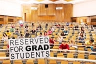 A lecture hall of students with two empty rows of chairs at the bottom and a sign that says "reserved for grad students."