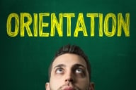 A male student looks up at the word "orientation" written in yellow on a green chalkboard behind him.