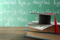 A stack of books, topped by cash and a graduation cap, in front of a chalkboard.