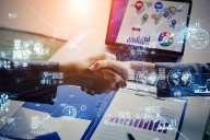 Two male hands shake while there are blue "digital" icons floating around. A computer screen and paper bar chart are in the background.