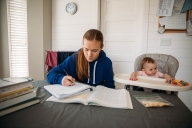 A mother sits with her baby daughter, who is in a high chair at the kitchen table while her mother studies for a university class.