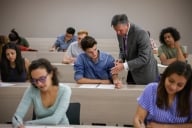 A professor speaks to a student in his lecture hall class, as other students are working on assignments.