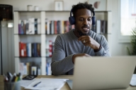 A man with gray in his beard sits at a desk pointing at a laptop with a bookshelf behind him.