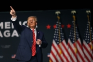 Donald Trump, in a blue suit and red tie and standing in front of American flags, points up.