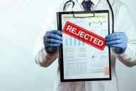 A man wearing a lab coat and a gloves holds up a paper covered in words and graphs but blocked by the word "rejected" in red.