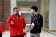 A man in a red shirt talks to a man in a black shirt in a high school hallway