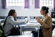 A student and professor sit at opposite sides of a classroom desk, with laptop open.