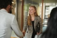 A young redheaded woman in professional attire shakes hands with a man whose back is turned to the camera (photo).