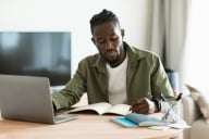 Black male college student sits at his laptop taking notes at home as he listens to a webinar.