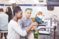 Two college students work together in the chemistry lab. She is carefully measuring solutions into two different beakers. He is watching closely. Other students are working in the background.