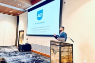A man with a beard stands at a podium in front of a PowerPoint with the JED logo on it.