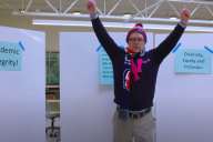 A man stands in front of whiteboards with signs taped to them reading "academic integrity" and "diversity, equity and inclusion" with both hands raised excitedly.