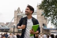 Portrait of a happy student in London holding a notebook and smiling near Tower Bridge