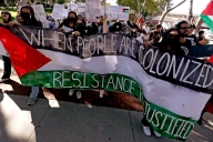 Student protesters have unfurled a large banner that features the Palestinian flag's colors and reads “When people are colonized, resistance is justified.” More protesters can be seen in the background, some holding signs that say things like “from the river to the sea, Palestine will be free” and “resistance against colonization is a human right.”