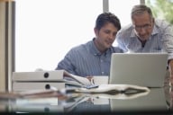 Older man leans over younger student who is looking at a computer and seems to be giving him advice