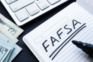 The word "FAFSA" is written in black marker on a page in an open notebook, which lies next to a stack of U.S. currency and the corner of a laptop keyboard.