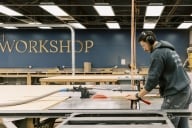 A man practices carpentry with the word "WORKSHOP" emblazoned on the wall behind him. 
