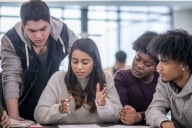 Four students of various ethnicities gather in a classroom, a woman gesturing as she explains something and two seated male students and one standing male student look on.