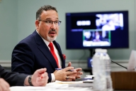 Education Secretary Miguel Cardona, in a blue jacket and red tie, sits with his hands on a table in a green room.