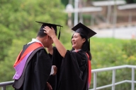 A student wearing a graduation robe and cape fixes a peer's graduation cap