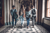 A group of four students, carrying backpacks, walk away from the camera down the hallway of a university building.