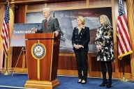 Republican senator Bill Cassidy stands a podium next to Senators Joni Ernst and Shelley Moore Capito.