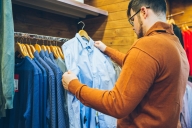 A man in a yellow-orange shirt holds up a blue button-down shirt while shopping in a clothing store. 