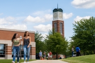 Two girls walk on a sidewalk with a college campus behind them. 