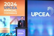A man is on a stage standing behind a podium. There is a background behind him with “2024 UPCEA” emblazoned on blue and orange. 