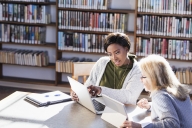 Two women working together in a library at a table. One has a laptop and the other is using a digital tablet to share information.