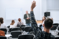 A young man raises his hand in the back of the classroom. In the background, out of focus, a professor stands at the front of the classroom.