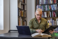 A man with gray hair and glasses sits at a table with laptop and book open, holding a clipboard, with filled bookshelves behind him.