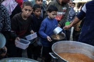 Displaced Palestinian children gather around a vat of what appears to be soup, holding containers to collect food aid.