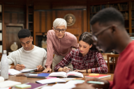 A group of three students studying at a table with the help of a professor standing behind them.