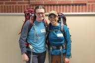 Two students wearing blue shirts and backpacks smile for a photo in front of a brick wall