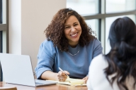 A mature woman smiles at an unrecognizable female student while writing on a notepad