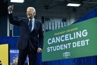 President stands in a suit jacket and tie holding a thumb up. A sign saying, “Canceling Student Debt,” is displayed behind him.