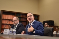 Two men in suits sit at a table with microphones