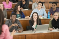 A multiethnic group of college-aged students sit in a row in a lecture hall listening to and apparently enjoying their professor whose back is turned to the camera.