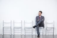 Man in suit sits alone impatiently waiting surrounded by a group of empty chairs