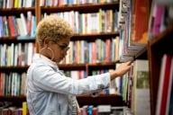 A student with short curly blonde hair, glasses and hoop earrings wearing a denim jacket looks for book on a bookshelf.