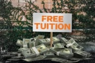 A sign reading “Free Tuition” is situation on a pile of money in front of an ivy-clad wall.